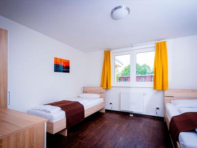 Ein einfaches Hotelzimmer mit zwei Einzelbetten, Holzfußboden und gelben Vorhängen am Fenster.