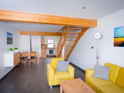 Wohnzimmer mit gelbem Sofa, Holztisch und offener Küche unter einer Holztreppe in einer Ferienwohnung.