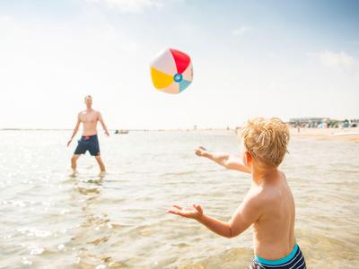 Ein Kind spielt mit einem Erwachsenen am flachen, sonnigen Strand mit einem bunten Strandball.