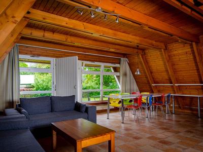 Gemütlicher Dachbodenraum mit Holzbalken, Sofa, Tischen und großen Fenstern mit Blick ins Grüne.