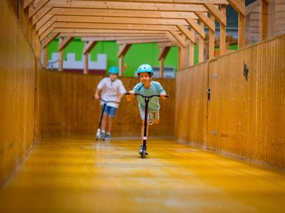 Kind mit Helm fährt Roller in einem langen, hölzernen Flur, erwachsener Mann joggt dahinter