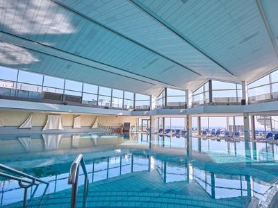 Helles Indoor-Schwimmbad mit großen Fenstern, blauer Wasserfläche und Liegestühlen am Rand.