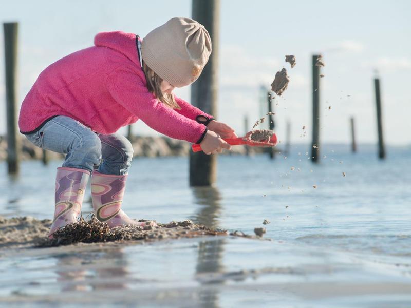 Ein Kind in rosa Jacke und Gummistiefeln spielt am Wasser und wirft Sand in die Luft.