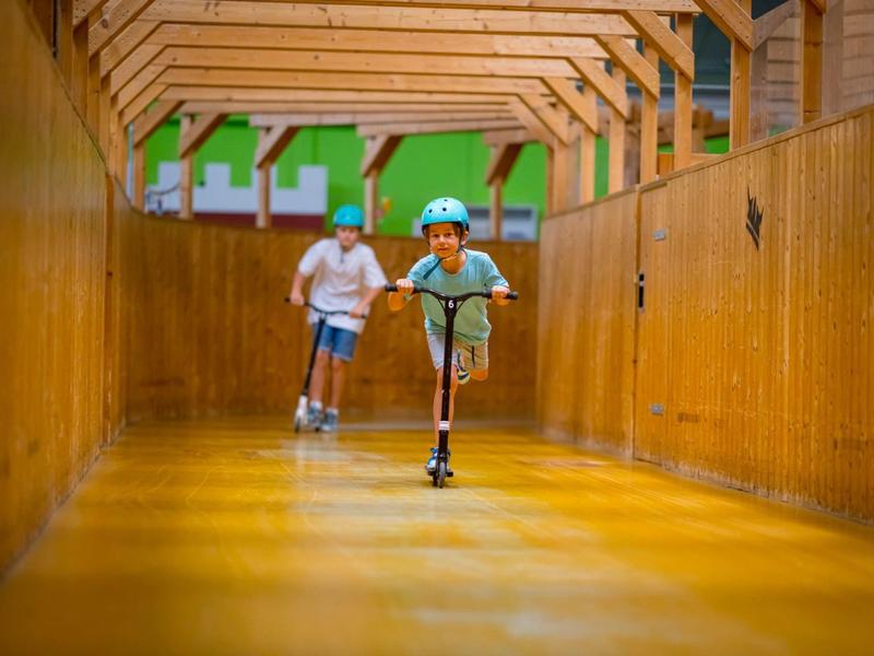 Kind mit Helm fährt Roller in einem langen, hölzernen Flur, erwachsener Mann joggt dahinter