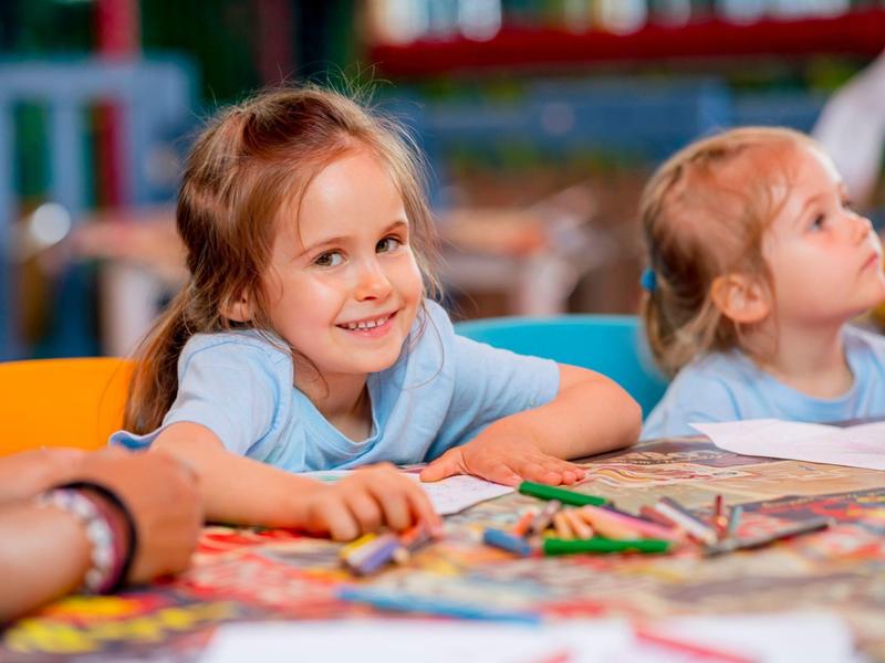 Zwei kleine Mädchen sitzen an einem Tisch voller Buntstifte und zeichnen fröhlich.