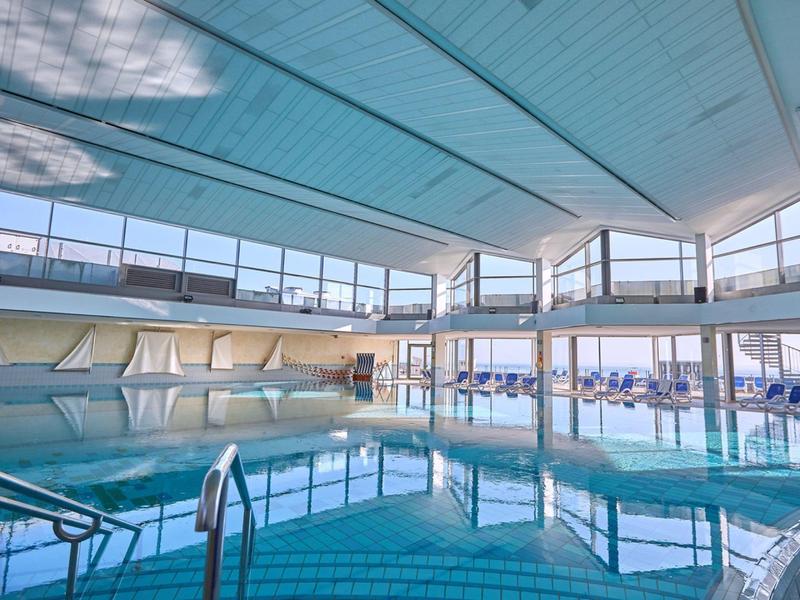 Helles Indoor-Schwimmbad mit großen Fenstern, blauer Wasserfläche und Liegestühlen am Rand.