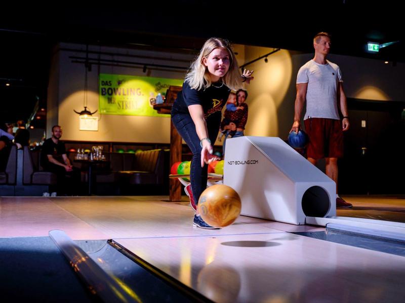 Junge Frau wirft Bowlingkugel auf Bahn in Bowlinghalle mit anderen Spielern im Hintergrund.