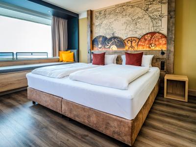 Helles Hotelzimmer mit großem Doppelbett, moderner Einrichtung und Sitzbereich am Fenster.