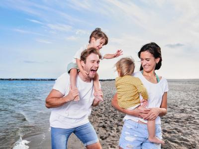Familie mit zwei Kindern am Strand bei Sonnenuntergang, Mutter hält Kind im Arm, Vater trägt Kind auf Schultern.