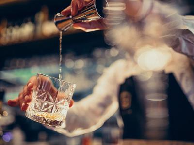 Eine Hand gießt ein Getränk aus einem Metallbehälter in ein Glas mit Eiswürfeln in einer Bar.