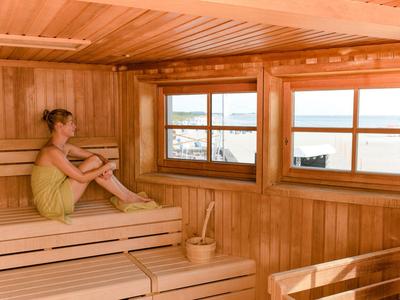 Žena v žlutém ručníku sedí uvolněně v dřevěné sauně s okny s výhledem na moře.