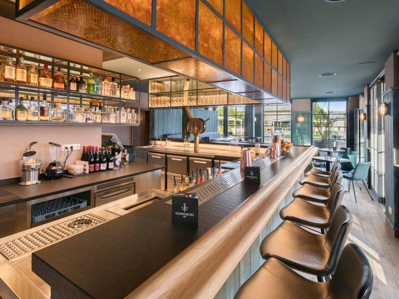 Moderní hotelový bar s dlouhými pulty, barovými židlemi a velkými okny s výhledem ven.