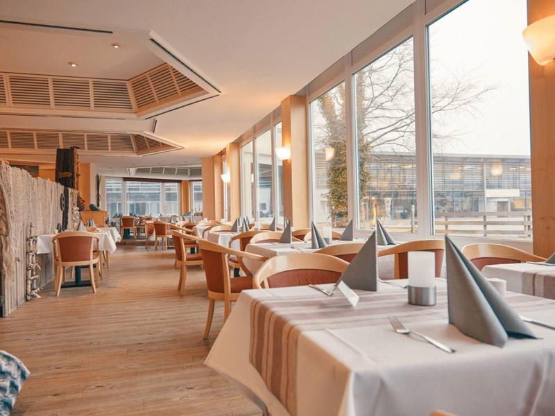 Světla restaurace s dřevěnou podlahou, velkými okny a prostřenými stoly se šedými ubrousky.