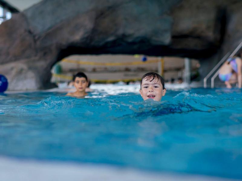 Dva chlapci plavou v krytém bazénu s kamennou dekorací v pozadí.
