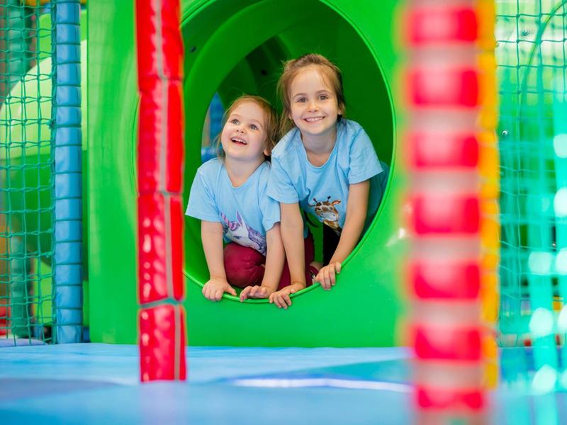 Dvě usměvavé děti si hrají v barevném vnitřním hřišti s tunelem a horolezeckými prvky.