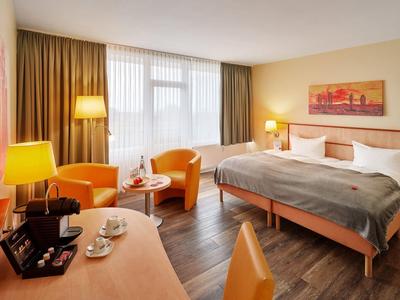 Modern hotel room with double bed, two chairs, desk, and large windows with curtains.