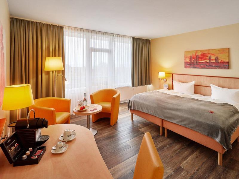 Modern hotel room with double bed, two chairs, desk, and large windows with curtains.