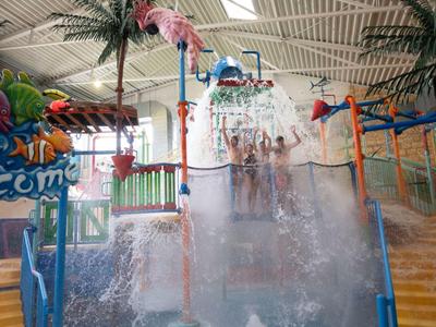Wasserspielplatz mit Wasserfontänen, Rutschen und tropischen Pflanzen in einer Halle.