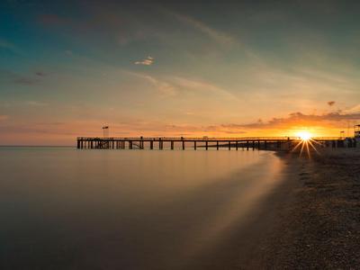 Langer Steg ragt bei Sonnenuntergang über ruhiges Meer an einem Sandstrand.