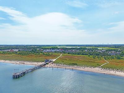 Langer Holzsteg führt über ruhiges Wasser zu einem Sandstrand mit Dünen und Wiesen bei sonnigem Himmel.