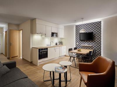 Modernes Wohnzimmer mit Küchenzeile, Esstisch, Sofa und Sessel in warmen Tönen.