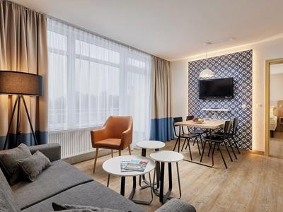 Modern eingerichtetes Hotelzimmer mit Sofa, Sessel, Esstisch und Flachbildfernseher an der Wand.