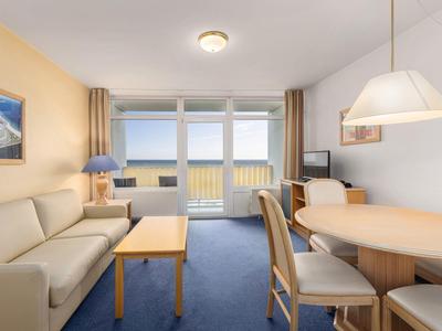 Jasny salon z sofą, stołem jadalnym i dużymi oknami z widokiem na morze.