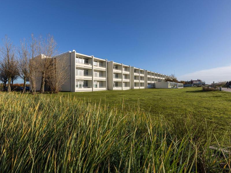 Nowoczesny budynek hotelowy z balkonami, otoczony dużym zielonym trawnikiem i czystym niebieskim niebem.