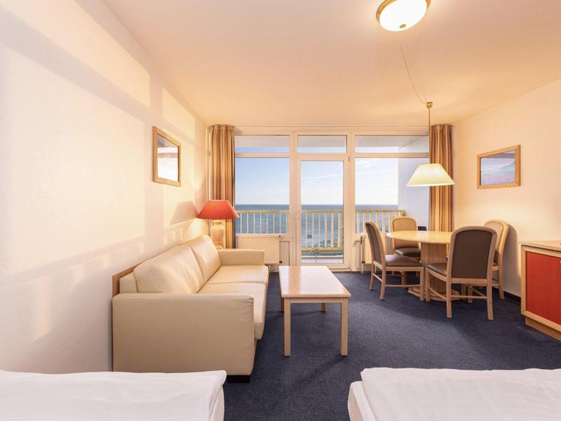 Jasny pokój hotelowy z balkonem, sofą, stołem jadalnym, krzesłami i widokiem na morze o zachodzie słońca.