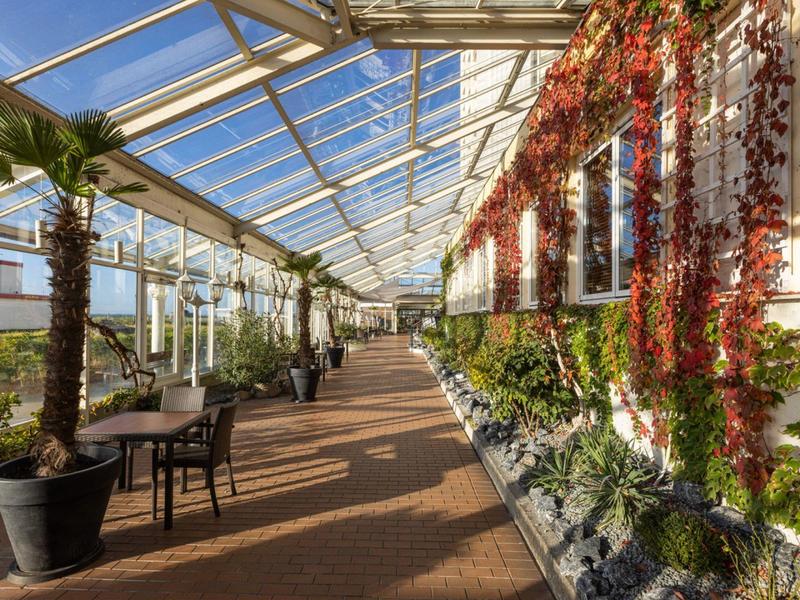 Jasna oranżeria ze szklanym dachem, roślinami i miejscami do siedzenia wzdłuż słonecznego korytarza.