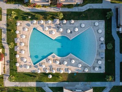 Drohnenaufnahme eines rechteckigen Hotelpools mit weißen Liegen und Sonnenschirmen im Außenbereich.