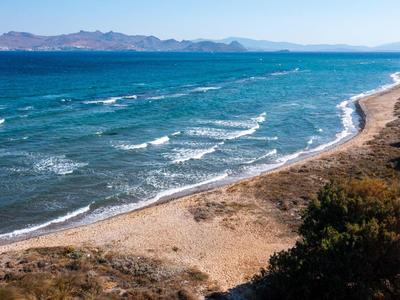 Strand mit sanften Wellen und Bergen im Hintergrund, bewachsen mit Büschen und Bäumen.