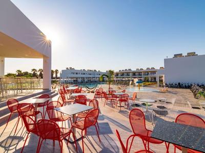 Terrasse mit roten Stühlen und Tischen vor einem modernen Hotelgebäude bei Sonnenschein.