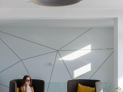 Moderner Raum mit geometrischer Wand, zwei Sesseln, Kissen und einem kleinen Tisch, Sonnenlicht fällt herein.