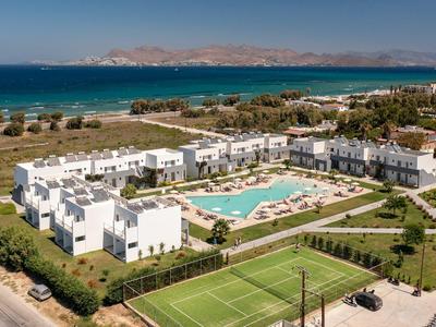 Große Ferienanlage mit weißen Gebäuden, Pool, Tennisplatz und Meer im Hintergrund bei klarem Himmel.
