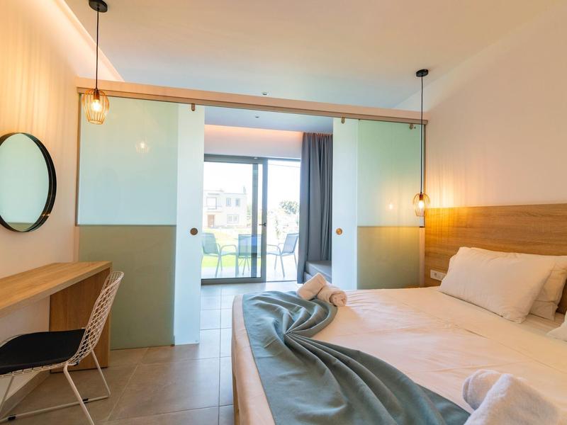 Bright hotel room with double bed, desk, chair, and balcony with view.