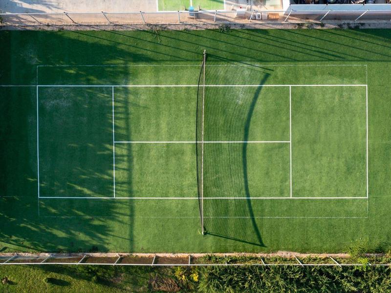 Drone view of a green tennis court with court markings surrounded by fences.
