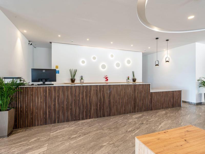 Modern hotel reception with wooden counter, plants, and bright lighting.
