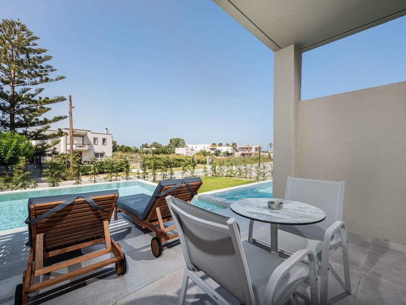 Balcony with table and chairs overlooking wooden loungers and a pool under a clear blue sky.