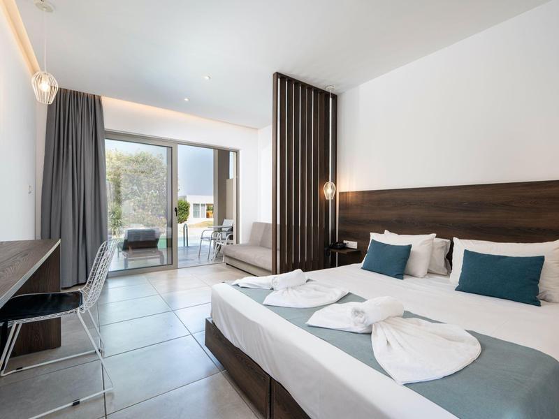 Modern hotel room with double bed, desk, chair, and balcony access overlooking greenery.