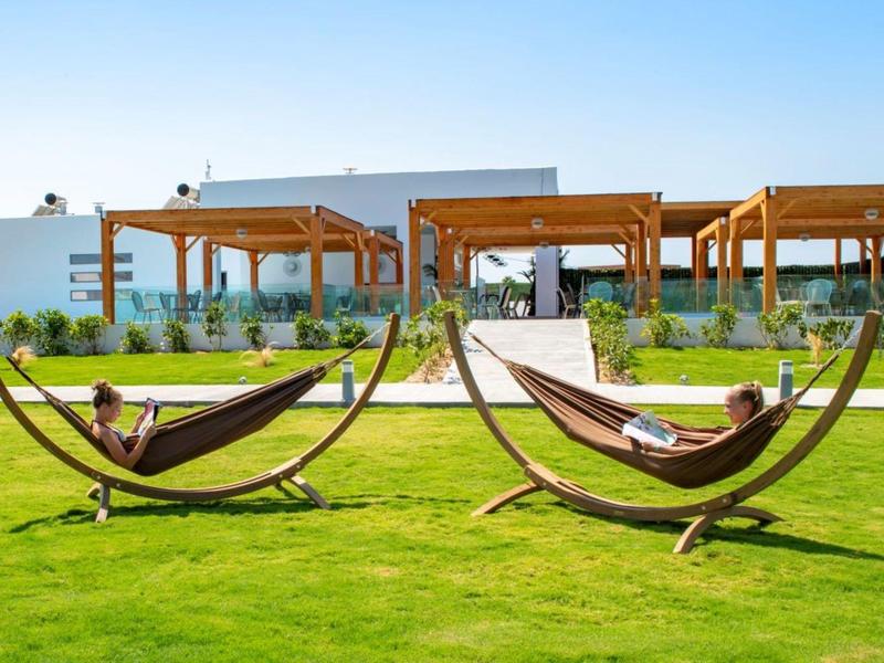Two people relax in hammocks on a green lawn in front of a modern hotel building with pergolas.