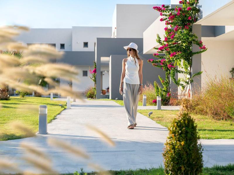 Woman in casual clothes walking on a path through modern hotel grounds with white buildings and garden.