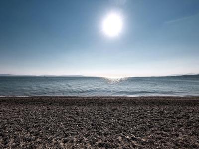 Strand mit dunkelgrauem Kies, ruhiges Meer und klarer Himmel mit strahlender Sonne.
