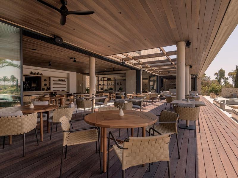 Moderner Außenbereich eines Restaurants mit Holzboden, runden Tischen und geflochtenen Stühlen.