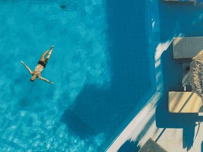 Person entspannt beim Schwimmen in einem blauen Pool neben Liegestühlen und Sonnenschirm.