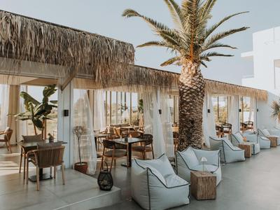 Outdoor-Sitzbereich eines Hotels mit Tischen, Stühlen und Palmen unter einem strohgedeckten Dach.