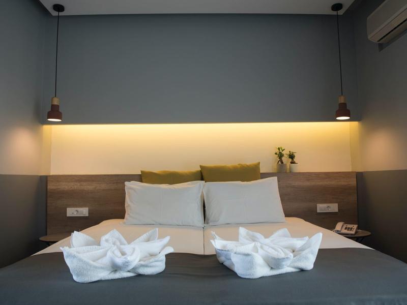 Jednoduše zařízený hotelový pokoj s manželskou postelí, dekoračními ručníky a nepřímým osvětlením.