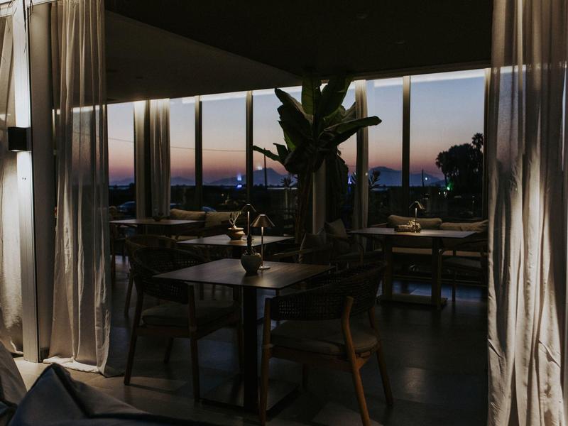 Elegantní restaurace s průhlednými závěsy a výhledem na západ slunce nad mořem.