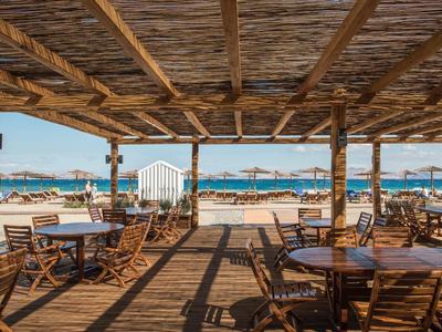 Offene Strandbar mit Holzdach, Tischen, Stühlen und Sonnenschirmen vor blauem Meer und Himmel.