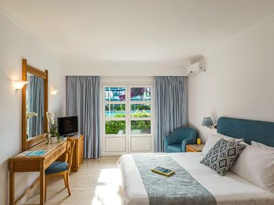 Helles Hotelzimmer mit Doppelbett, Schreibtisch, Sesseln und Blick auf einen Garten.